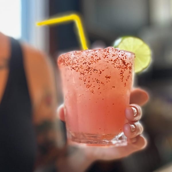 A hand holds a pink cocktail with a chili-salt rim, lime wedge, and yellow straw.