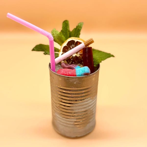 A cocktail in a tin can with a pink straw, dried orange, mint, and gummy worms.