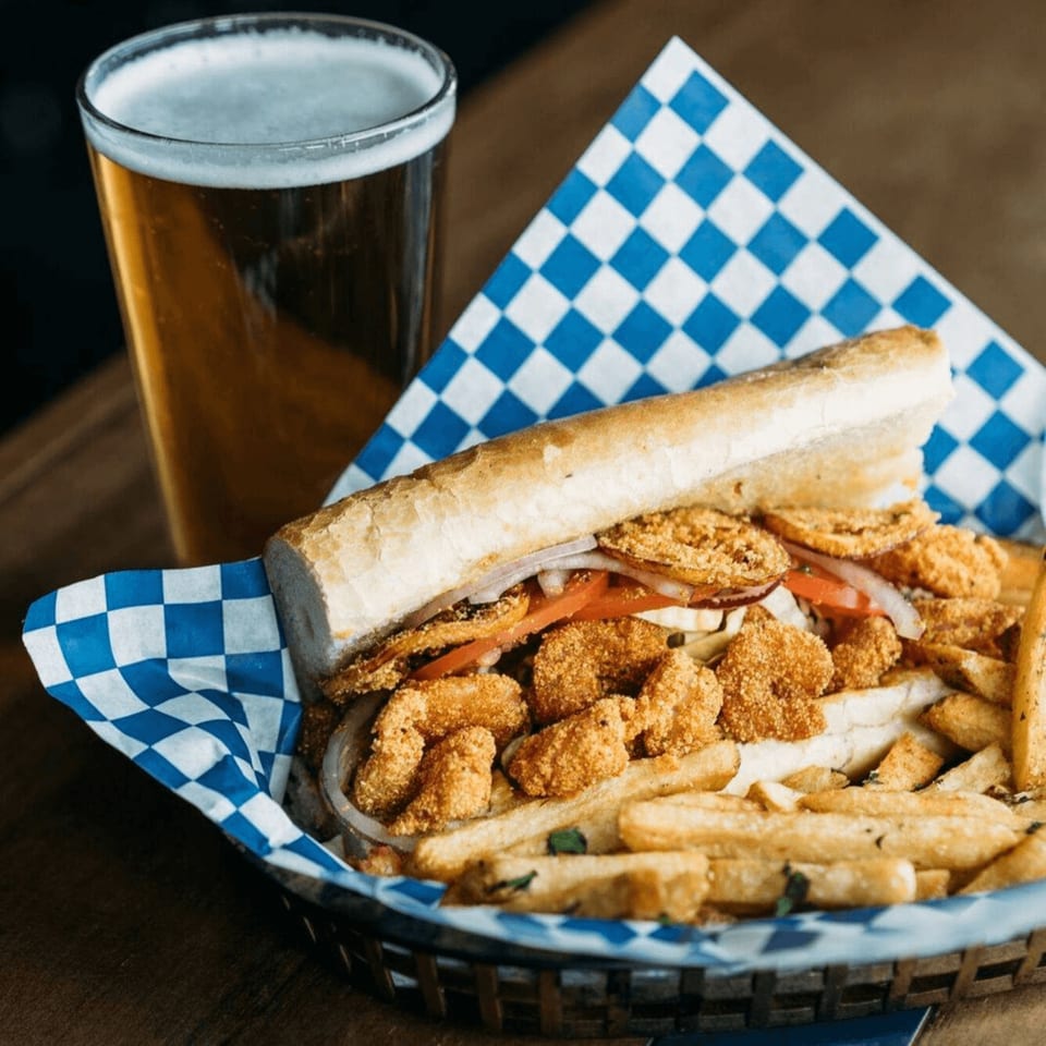 A fried shrimp po'boy with fries in a basket, alongside a glass of beer.