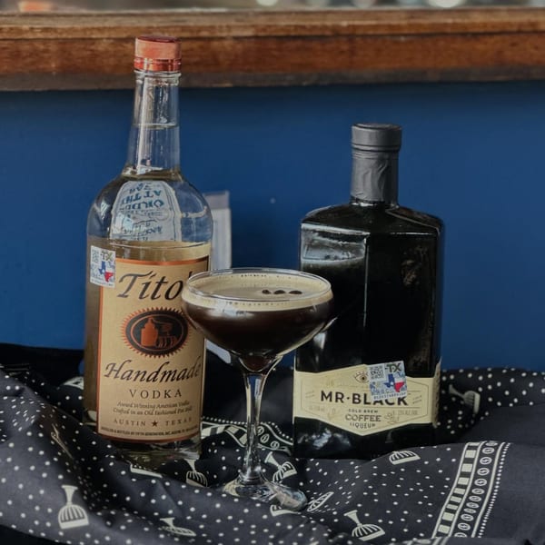 Espresso martini with coffee beans, Tito's Handmade Vodka, and Mr. Black Coffee Liqueur bottles on a patterned cloth.