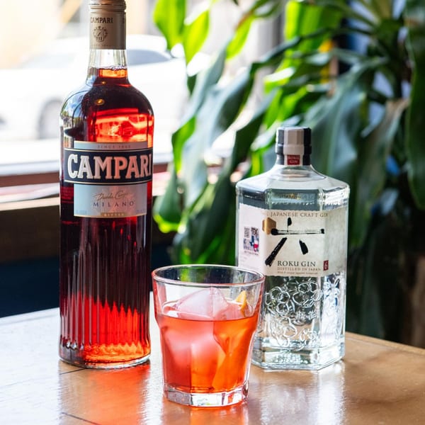 A Campari bottle, a Roku Gin bottle, and a Negroni cocktail on a wooden table.
