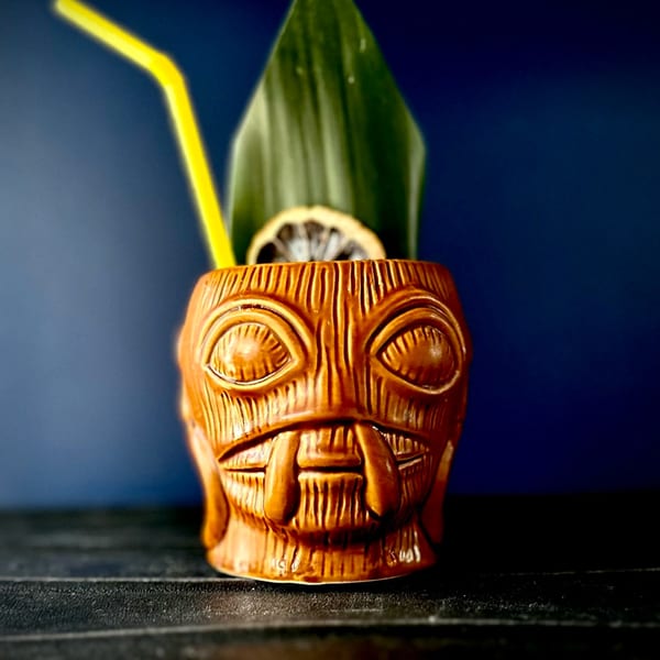 A tiki cocktail in a brown ceramic mug with a yellow straw, green leaf, and dried orange slice.