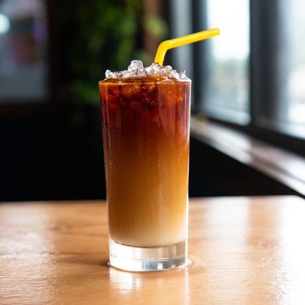 Iced coffee with a yellow straw, showing layers of dark coffee and light cream, on a wooden table.