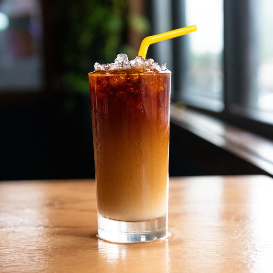 Iced coffee with a yellow straw, showing layers of dark coffee and light cream, on a wooden table.